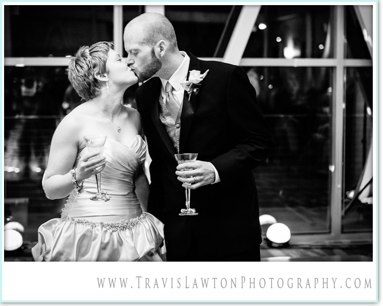 Husband and wife share a kiss as reception guests clink their glasses